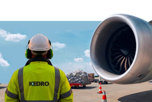 Aviation ground crew staff member standing next to an aeroplane engine turbine, waiting for takeoff. Wearing a higher visibility Kedro branded jacket, white hard hat, ear defenders