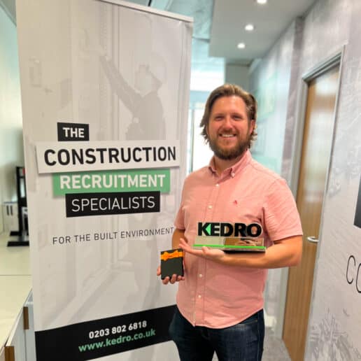 Bo Sparks holding a Kedro search employee of the month award and also standing next to a construction recruitment specialist's roller banner in the Kedro office in London.